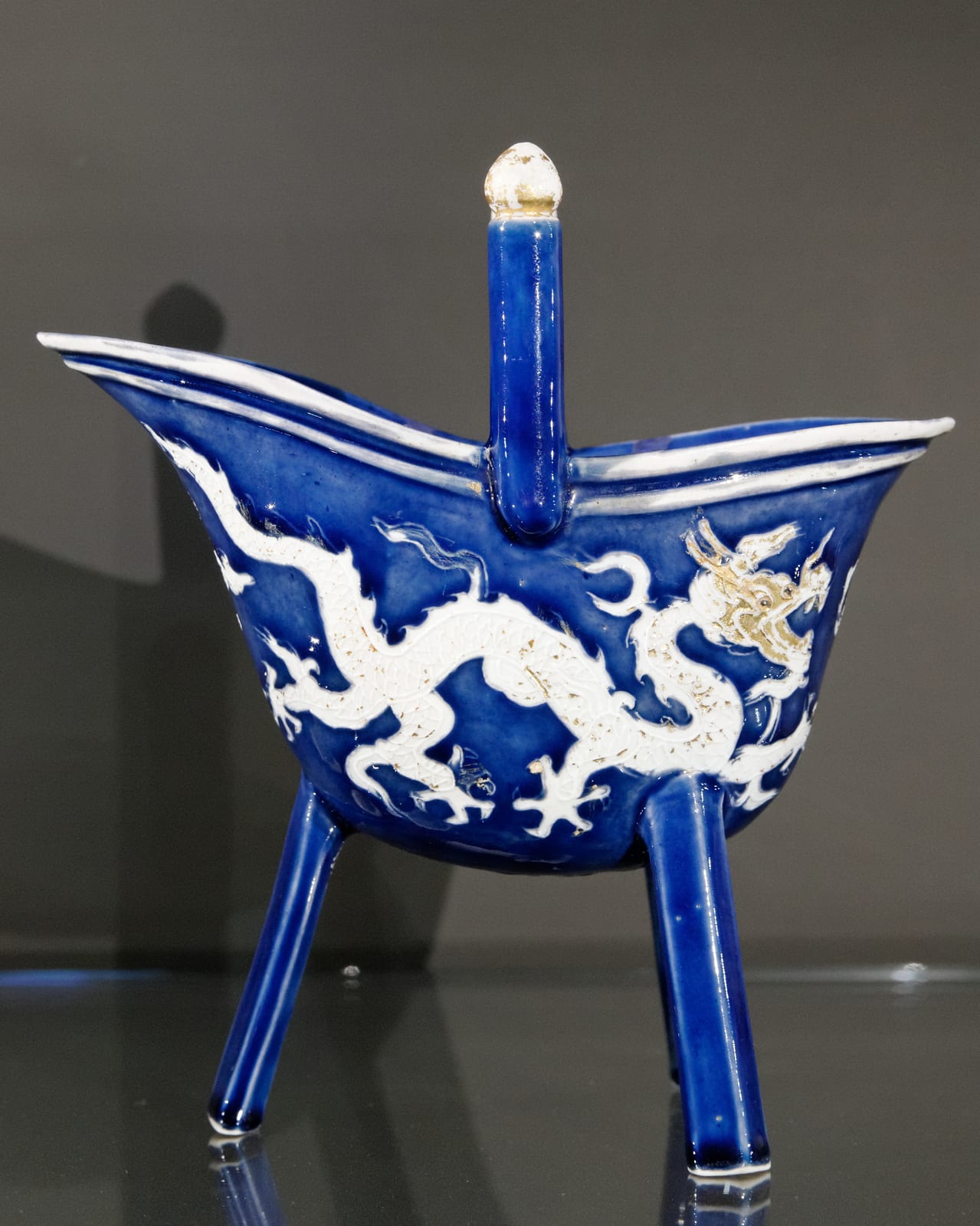 青い釉に白い龍文を入れた明代の磁器の爵。三本の脚と柱は残るが、注ぎ口と尾が短くなり、胴が丸い杯の形になっている